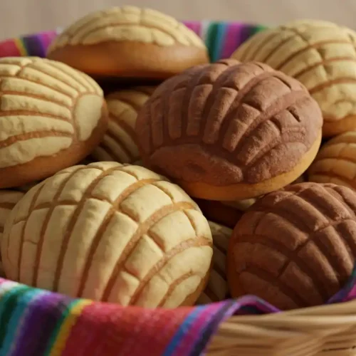 Fresh Mexican concha bread in vanilla and chocolate varieties arranged in a basket lined with colorful serape fabric, showing their distinctive shell-pattern tops