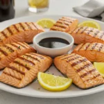 Grilled salmon fillets arranged around a dark dipping sauce, garnished with fresh lemon wedges on a white plate