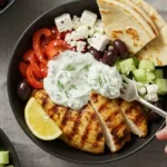 A plate of sheet pan chicken pitas with tzatziki sauce, olives, tomatoes, and cucumbers.