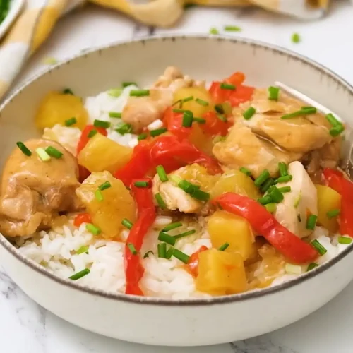 A bowl of Sweet Hawaiian Crockpot Chicken served over white rice, featuring tender chicken, red bell peppers, and pineapple chunks, garnished with fresh green onions.