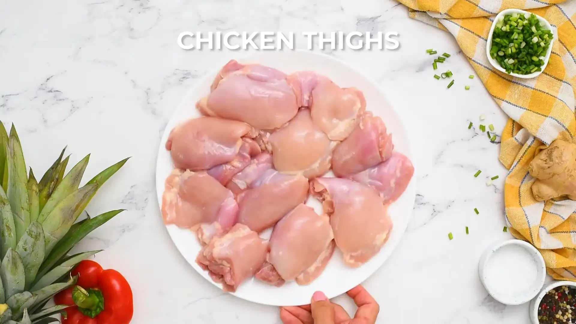 A plate of raw boneless, skinless chicken thighs ready for preparation, with "SALT" text overlay, set on a marble countertop with green onions and pineapple in the background.