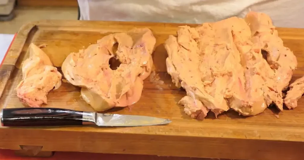 Fresh foie gras lobes being cleaned and deveined on a wooden cutting board with a paring knife.