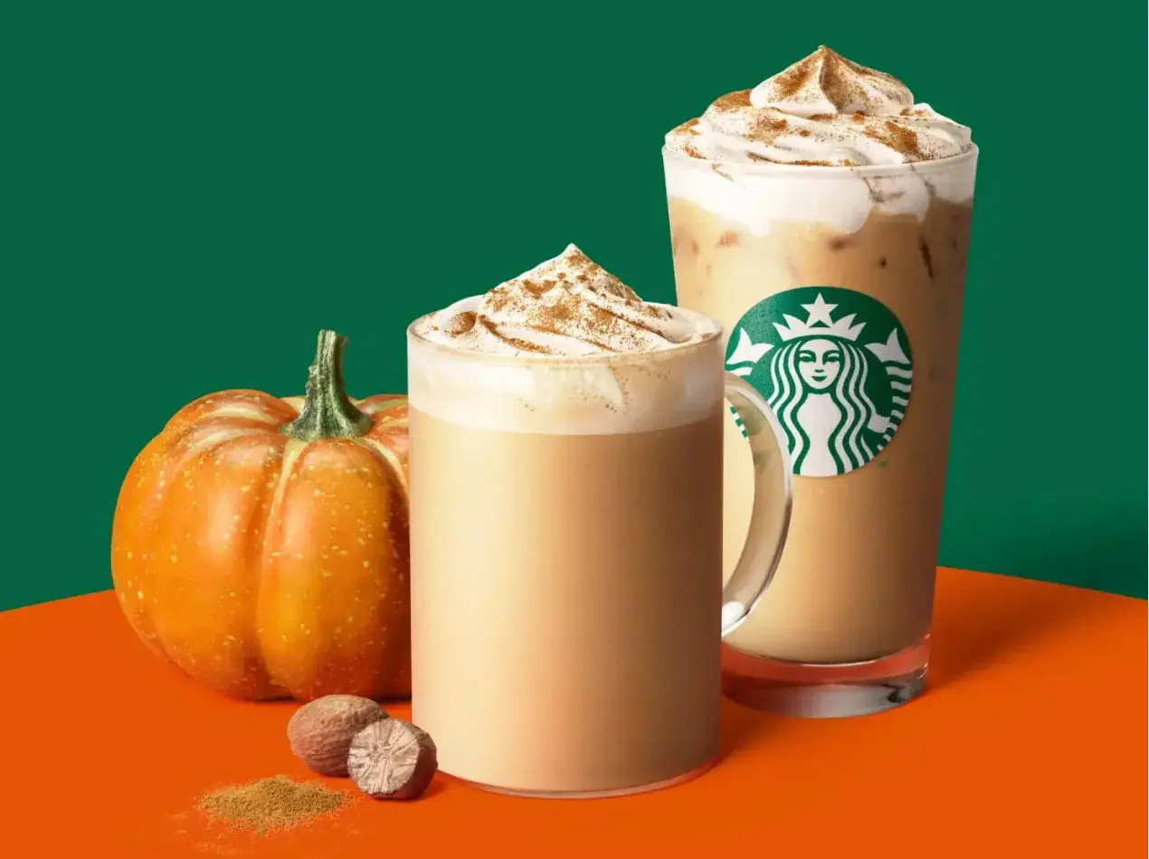 Two Starbucks Pumpkin Spice Latte Recipe drinks – one hot latte in a clear glass mug and one iced latte in a Starbucks cup, both topped with whipped cream and a dusting of pumpkin-pie spice, alongside a small pumpkin and whole nutmeg on an orange-and-green backdrop