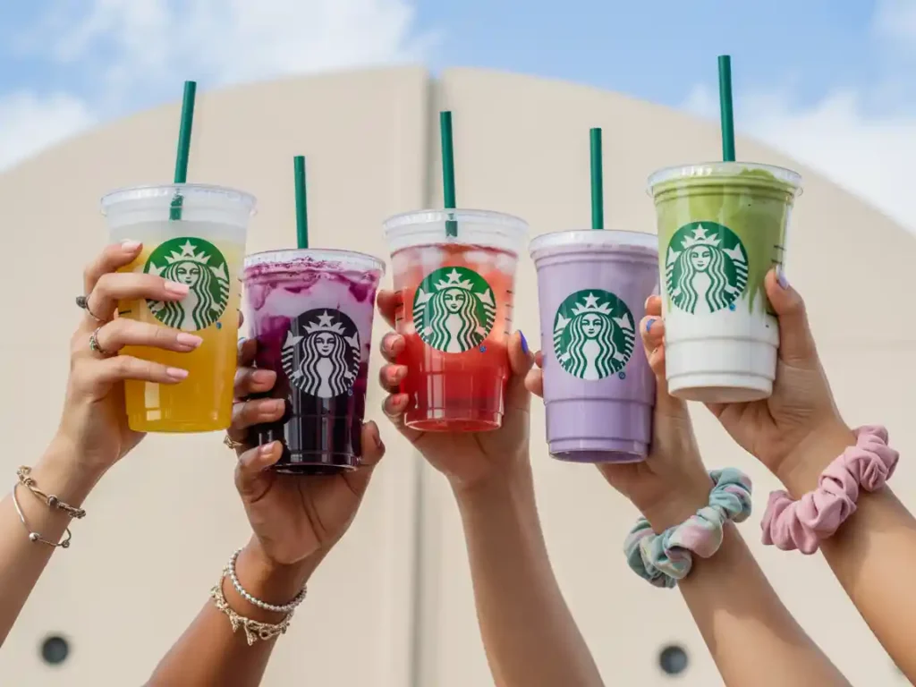 Hands holding colorful Starbucks Secret Menu drinks in yellow, purple, red, violet, and green against a bright sky background.